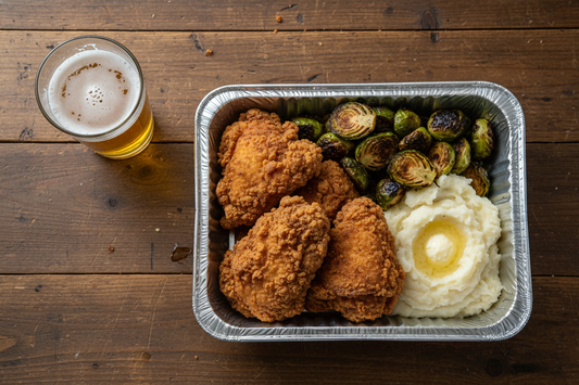 Crunchy southern fried chicken and brussel sprouts that are slightly chared on the grill with a side of creamy mashed potatoes in a foil takeout container (9x13 inch) size. on a kitchen table with a pint of beer 